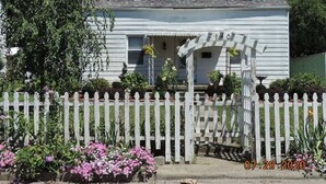 Une maison blanche à deux étages, avec une clôture en bois peinte en blanc, un porche abrité et des fleurs roses dans le jardin.
