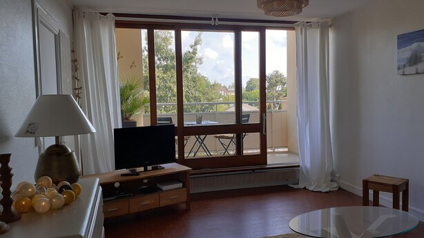 En stue med fjernsyn, et glasbord, en sofa og en balkon med bord og stole.