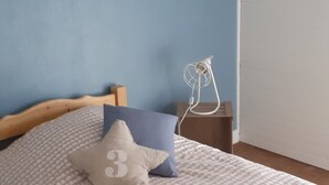 Et soveværelse med seng, puder, et natbord, en vægmonteret ventilator og et plakat.