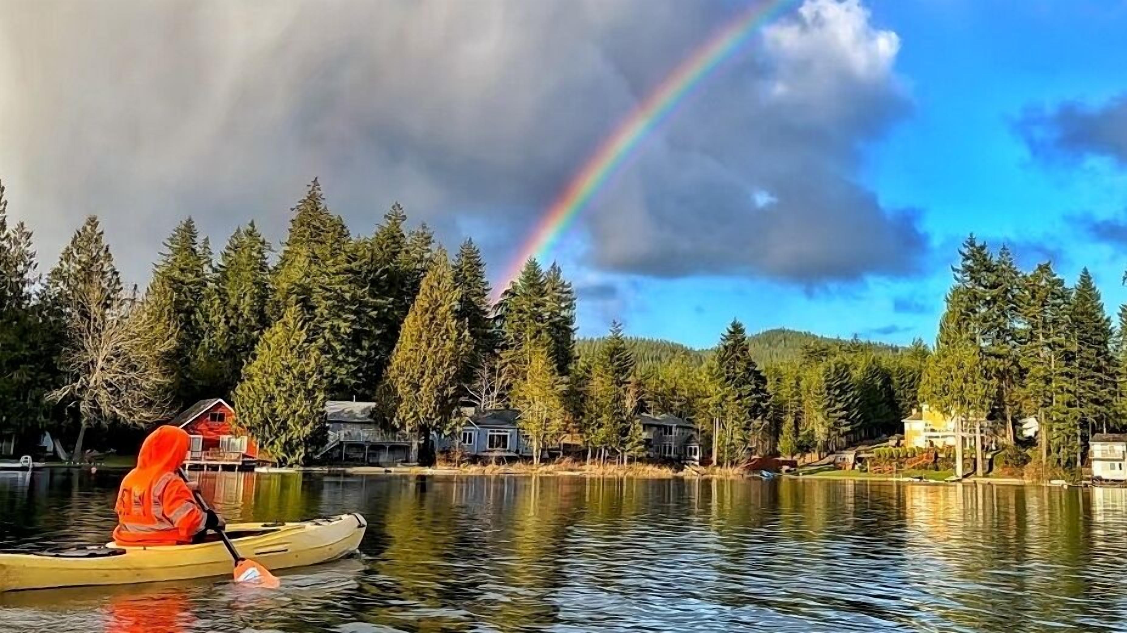 A person kayaking on a lake with houses and trees in the background, and a rainbow in the sky.