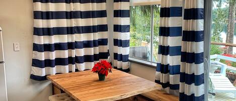 A wooden dining table with a bench, curtains, and a window with a view of greenery.
