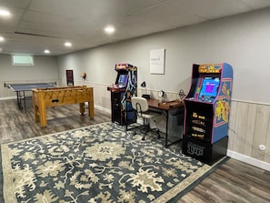 Quarto com mesa de pingue-pongue, mesa de futebol de mesa, duas máquinas de jogos eletrônicos, uma escrivaninha e um tapete estampado.