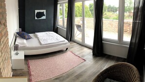 A bedroom with a bed, a small bedside table, a red rug, a wicker chair, and a view of the outdoors through large windows. - 5 minutes from the national park Holiday home for hikers, cyclists, anglers (Lunow-Stolzenhagen)