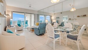 Un amplio salón con una mesa de comedor de cristal rodeada de sillas de mimbre blanco, un sillón azul y un sofá blanco.