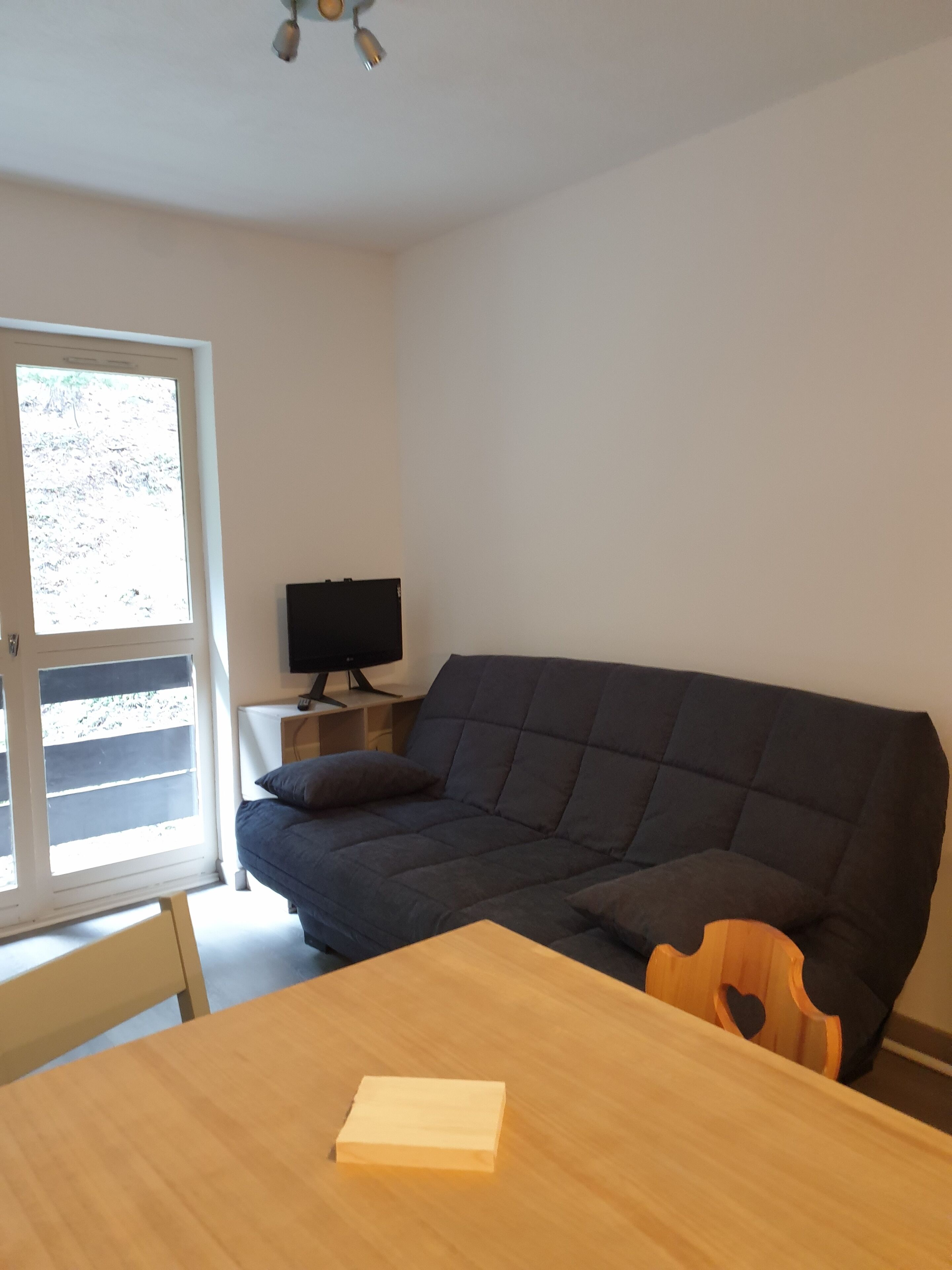 Een kamer met een donkere bank, een klein houten tafeltje, een televisie op een dressoir en een raam met jaloezieën.