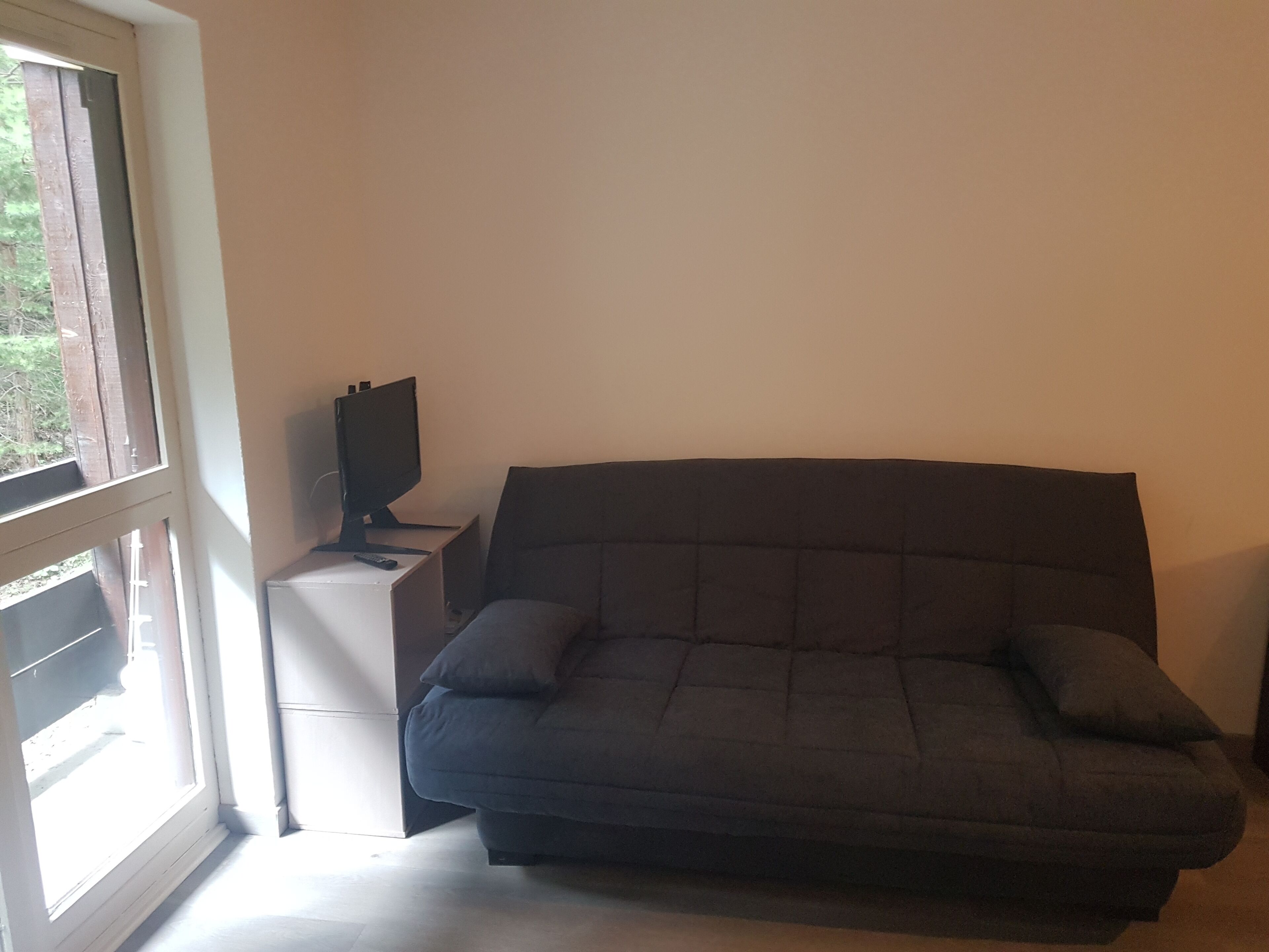 A room with a dark-colored sofa, a small white side table, a television on the table, and a window providing a view of greenery outside.