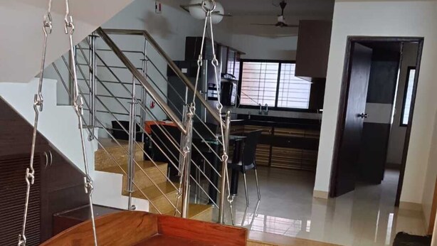 A modern interior with a wooden swing chair suspended from the ceiling, a staircase, and a kitchen area in the background.