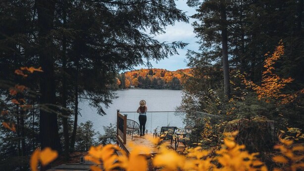 Eine Person steht auf einer Holzdielen-Terrasse mit Blick auf einen See, umgeben von Bäumen und Herbstlaub.