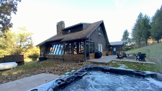 A hot tub in front of a rustic cabin with a stone chimney, large windows, and a patio area with outdoor seating. - Whispering Pines Retreat-Newly Renovated Countryside Home (Bedford)