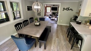 A dining area with a wooden table, black chairs, and a pendant light. There is a kitchen area with a countertop and stools. - Whispering Pines Retreat-Newly Renovated Countryside Home (Bedford)