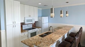 A modern kitchen with white cabinets, a granite countertop, a built-in oven, and a stainless steel sink. There are three pendant lights and a bar stool area with brown leather chairs. - Club Paradise Beach Condo-Top Floor, Ocean View, King Bed, Pet Friendly (Galveston)