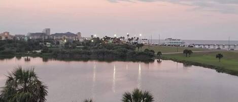 A calm lake with palm trees in the foreground, a pier extending into the water, and buildings in the background.