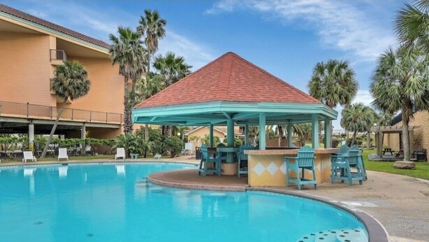 A swimming pool with a gazebo, lounge chairs, and palm trees. - Club Paradise Beach Condo-Top Floor, Ocean View, King Bed, Pet Friendly (Galveston)