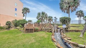 A landscaped area with a small waterfall, palm trees, and a wooden bridge. - Club Paradise Beach Condo-Top Floor, Ocean View, King Bed, Pet Friendly (Galveston)