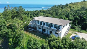 Un edificio moderno de dos plantas con techo plano, rodeado de abundante vegetación y vistas a una playa.