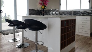 Una cocina moderna con una isla central, dos taburetes de bar, un jarrón con flores y vistas amplias a través de grandes ventanales.