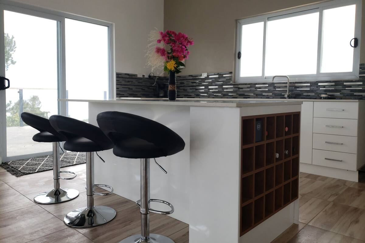 A modern kitchen with a central island, two bar stools, a vase with flowers, and a view through large windows.