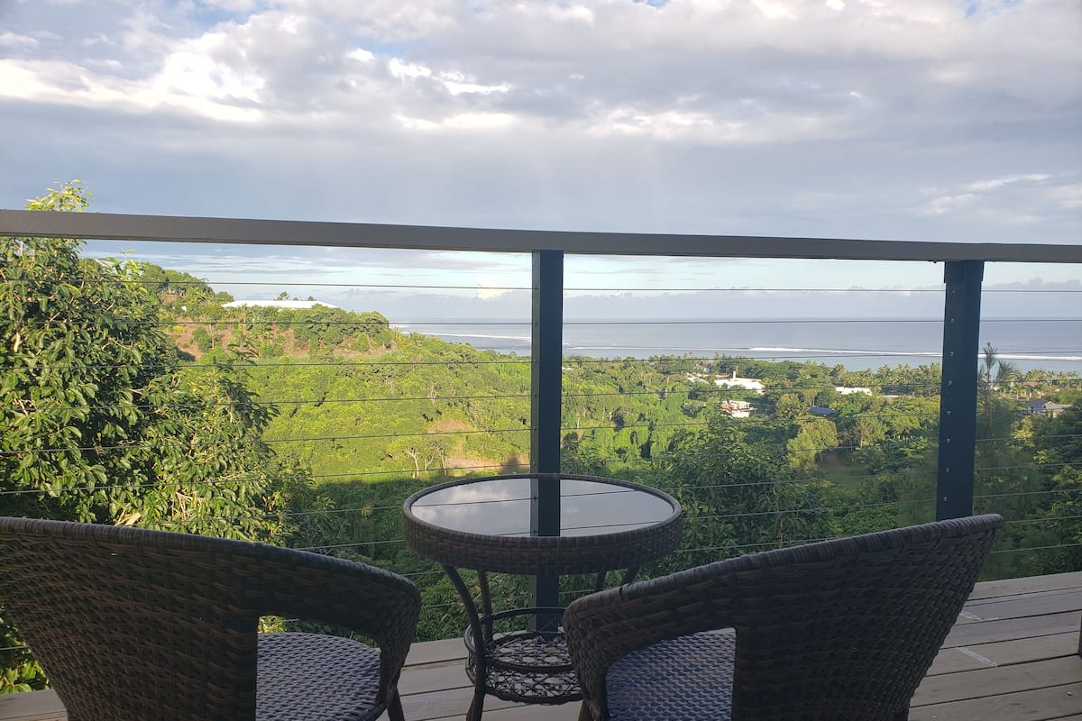 A balcony with wicker chairs and a small round table, overlooking a lush landscape with a view of the ocean and distant hills.