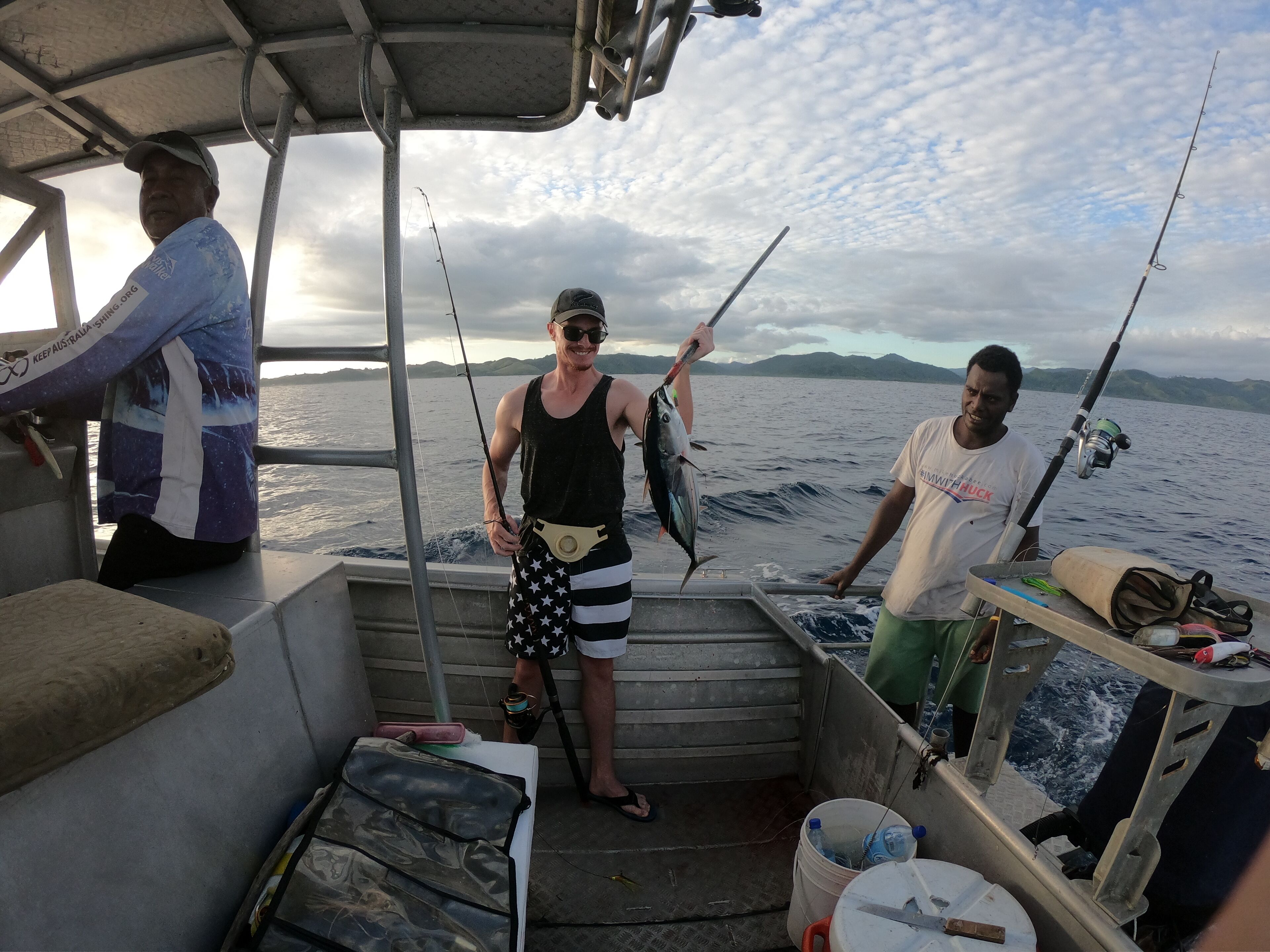 Interno di una barca con canne da pesca, una persona che tiene un pesce e una vista panoramica del mare e delle montagne sullo sfondo.