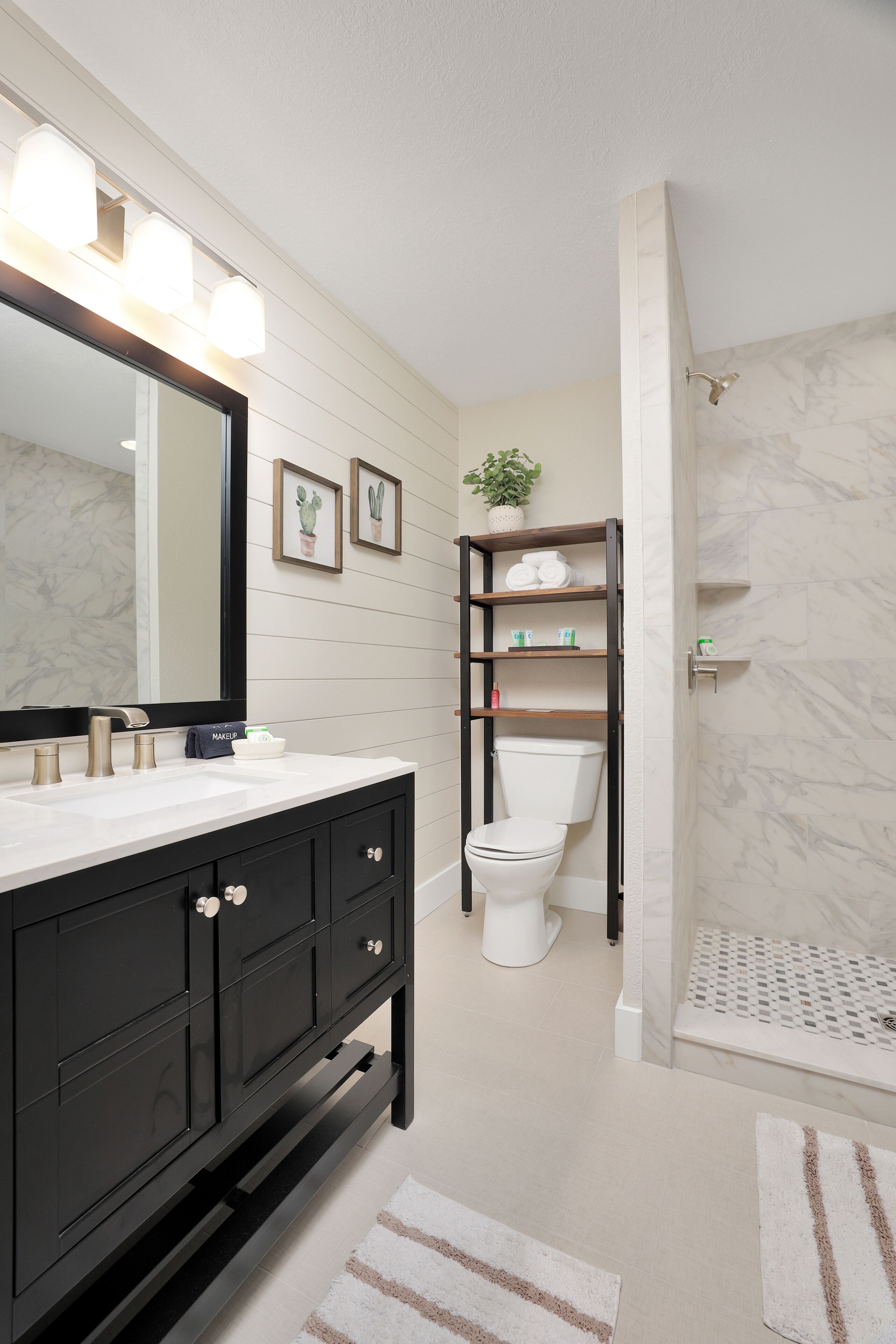 A bathroom with a black vanity, a white toilet, a shower with a marble wall, and a shelf with toiletries.