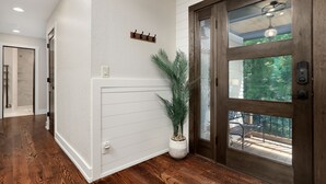 A hallway with wooden flooring, a white wainscoting wall, a potted plant, and a door with a glass panel and a balcony.