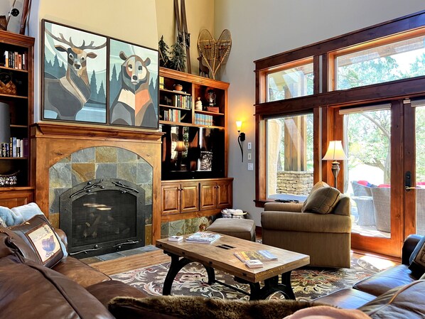 Uma sala de estar acolhedora com lareira, assentos confortáveis, estante de livros e uma grande janela com vista para o exterior.