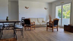 A living room with a sofa, two wooden chairs, a dining table with chairs, a vase, and a wooden chest of drawers. - Grand F3, Avec Balcon Plein sud Sans vis à vis (Vannes)