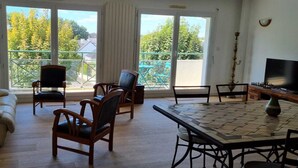 A living room with a sofa, chairs, a table, and a TV. There are large windows with a view of trees and a balcony. - Grand F3, Avec Balcon Plein sud Sans vis à vis (Vannes)