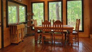 Interior de una cabaña de madera con una mesa de comedor y sillas, ventanas amplias con vistas al bosque y un estante con utensilios de cocina.