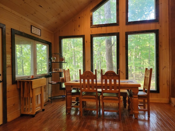 A wooden cabin interior with a dining table and chairs, large windows with a forest view, and a shelf with kitchenware.