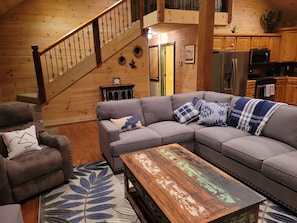 A cozy living room with a wooden staircase, a grey sofa, a brown leather armchair, and a rustic coffee table.