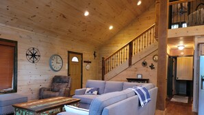 Interior acogedor de una cabaña de troncos, con una escalera de madera, un sofá azul grande, una mesa de centro rústica y un reloj de pared.