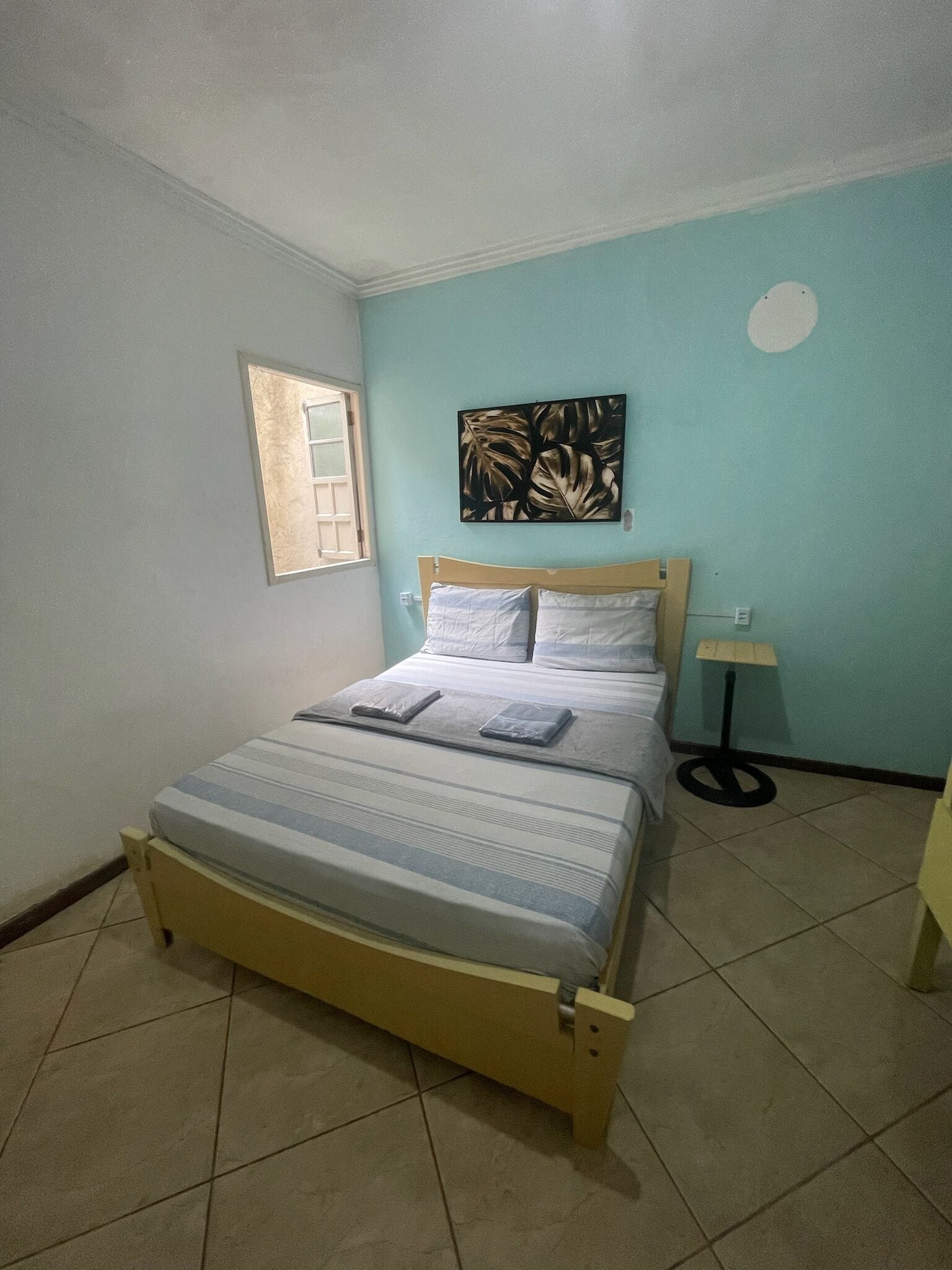 Un dormitorio con cama, ventana, una obra de arte en la pared y una mesita de noche con lámpara.