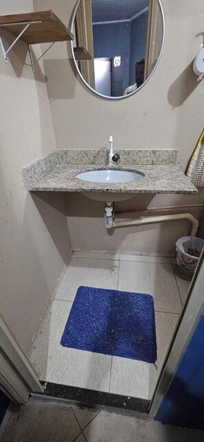 A bathroom with a granite countertop sink, a mirror, a blue mat on the floor, and a trash bin. - Hope Hostel (Guarapari)