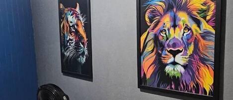 A room with two framed paintings of lions on the wall, a fan on a stool, and a patterned rug on the floor.
