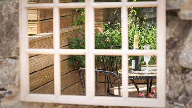 Un banco de madera y una mesa con una botella y un vaso, vistos a través de una ventana con entramado.