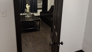 Una habitación con piso de madera, una puerta negra, un sofá oscuro, una mesita con una bandeja y una ventana con cortinas.