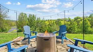 A stone patio with a fire pit, surrounded by blue Adirondack chairs, under string lights, with a view of a green landscape and trees. - Perfect for Family Reunions & Large Groups! 3 Pools - Games - Golf - Firepit! (Branson West)