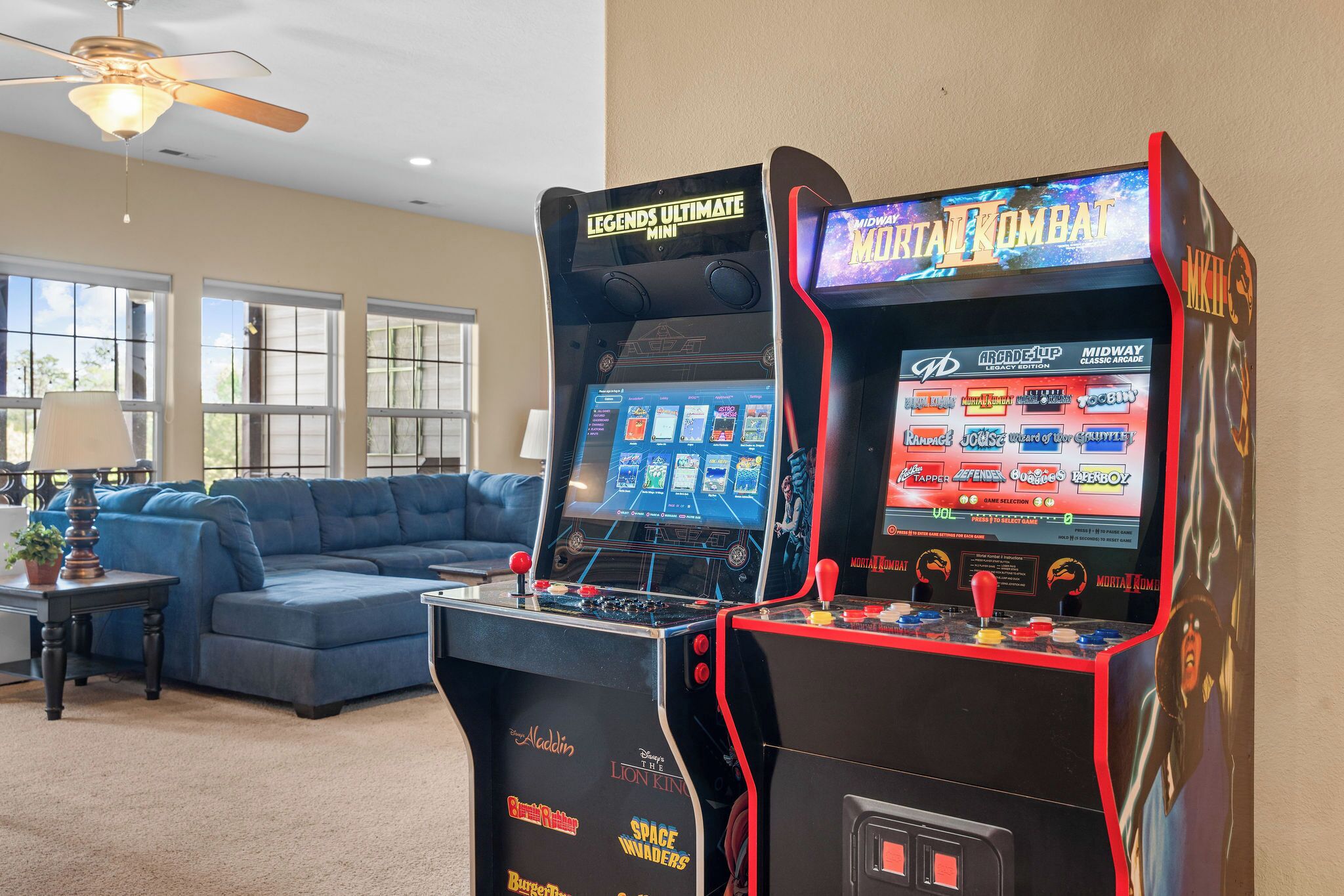 Two arcade machines in a living room with a blue sofa, a lamp, and a ceiling fan.