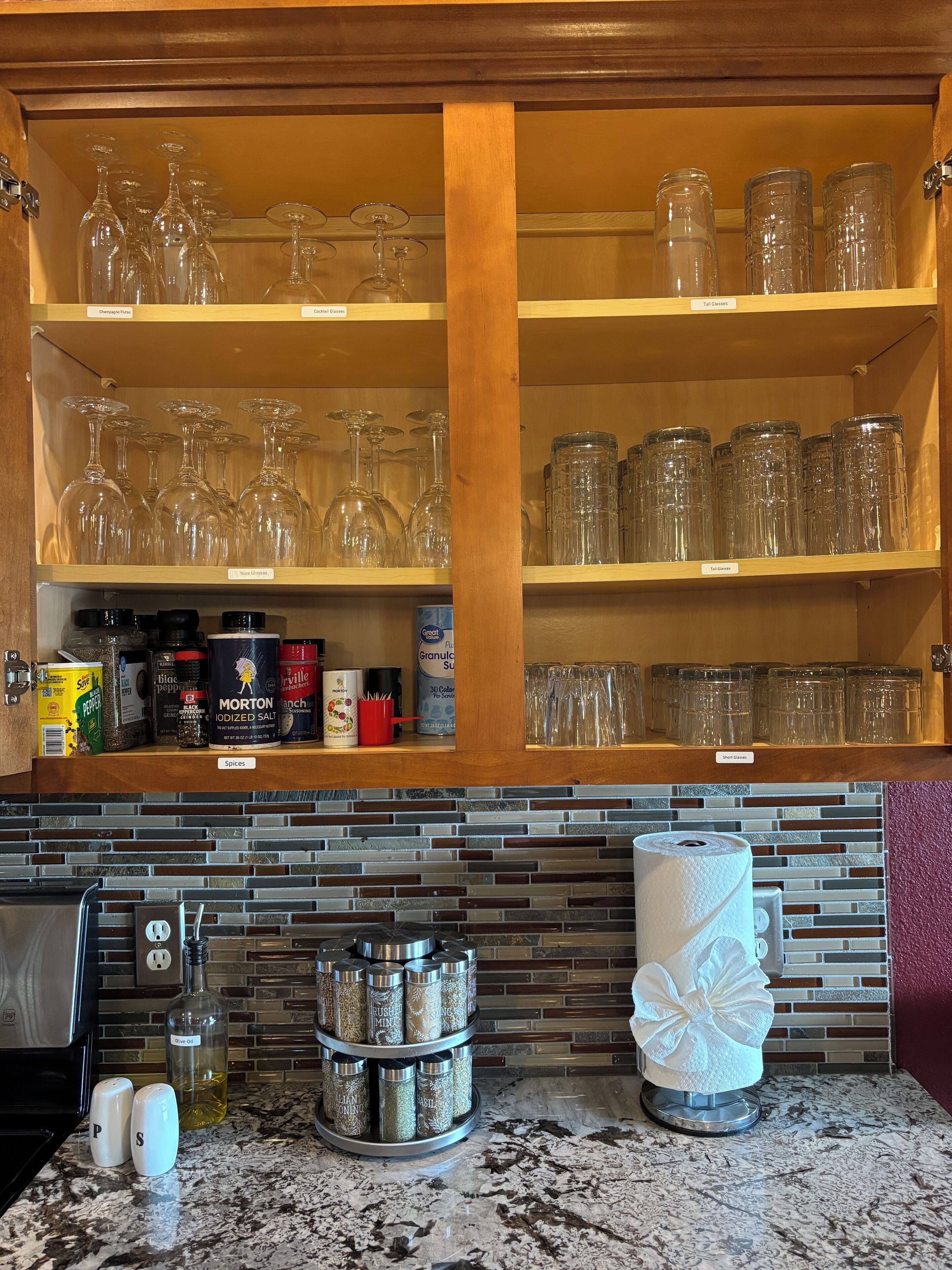 Una cocina con un estante de madera que sostiene copas de vino y frascos, un especiero y un dispensador de toallas de papel.