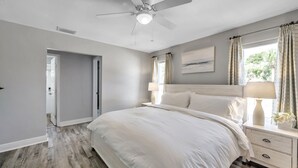 Un dormitorio con cama, mesita de noche, lámpara, ventilador de techo y ventana con cortinas.