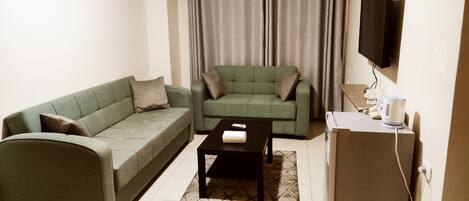 A living room with two green sofas, a coffee table, a flat-screen TV, and a refrigerator.