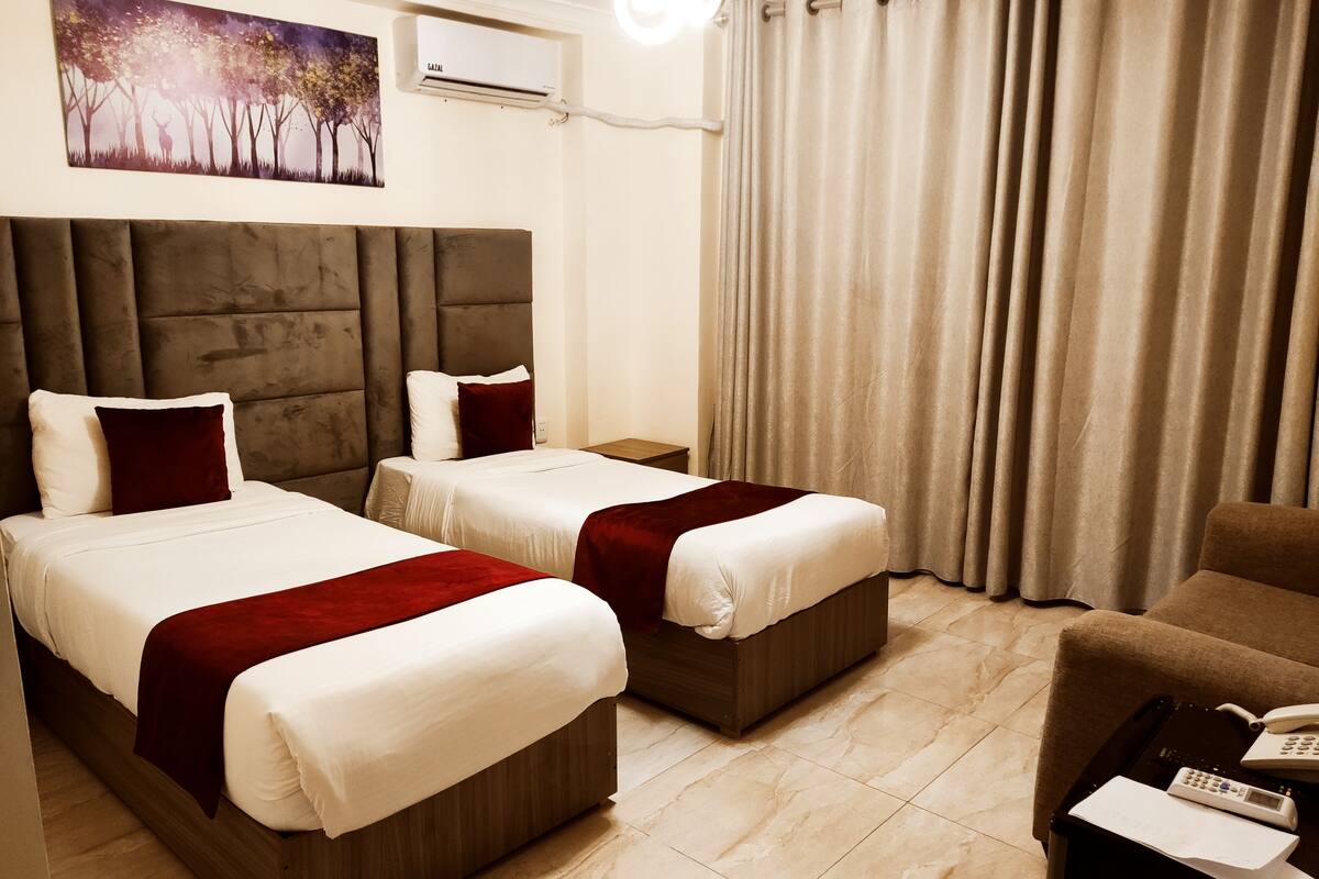 A hotel room with two beds, a headboard, a painting, an air conditioner, a curtain, a chair, and a table with a phone and papers.