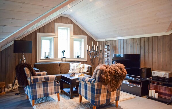 A cozy attic living room with a high ceiling, wooden walls, a large window, a chandelier, a television, and a bookshelf.