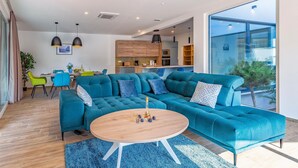 Un salon moderne avec un canapé d’angle turquoise, une table basse en bois et un coin repas comprenant une chaise bleue et une plante.