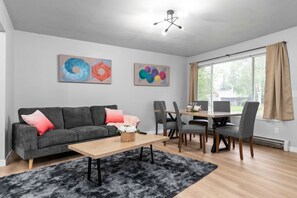 Een moderne woonkamer met een grijze bank, een houten salontafel en een eethoek met grijze stoelen en een houten tafel. Op de muur hangen abstracte schilderijen en er is een groot raam met gordijnen.