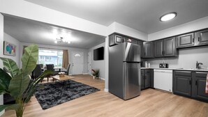 Une cuisine moderne avec des meubles gris, un réfrigérateur en acier inoxydable et un lave-vaisselle. On y trouve un coin repas avec une table et des chaises, ainsi qu’une grande plante au premier plan.