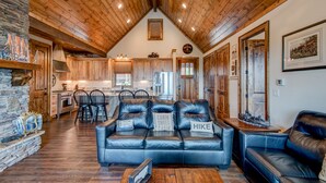 A spacious living room with a high wooden ceiling, a stone fireplace, and a modern kitchen in the background. - Lucky 7 at Eagles Nest: Long Range Views | Huge Deck | Firepit | Walk to Tennis! (Banner Elk)