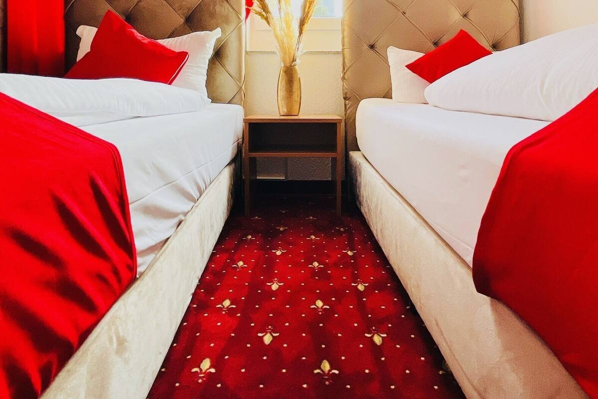 A hotel room with two beds, red curtains, a vase with dried flowers, and a red patterned carpet.
