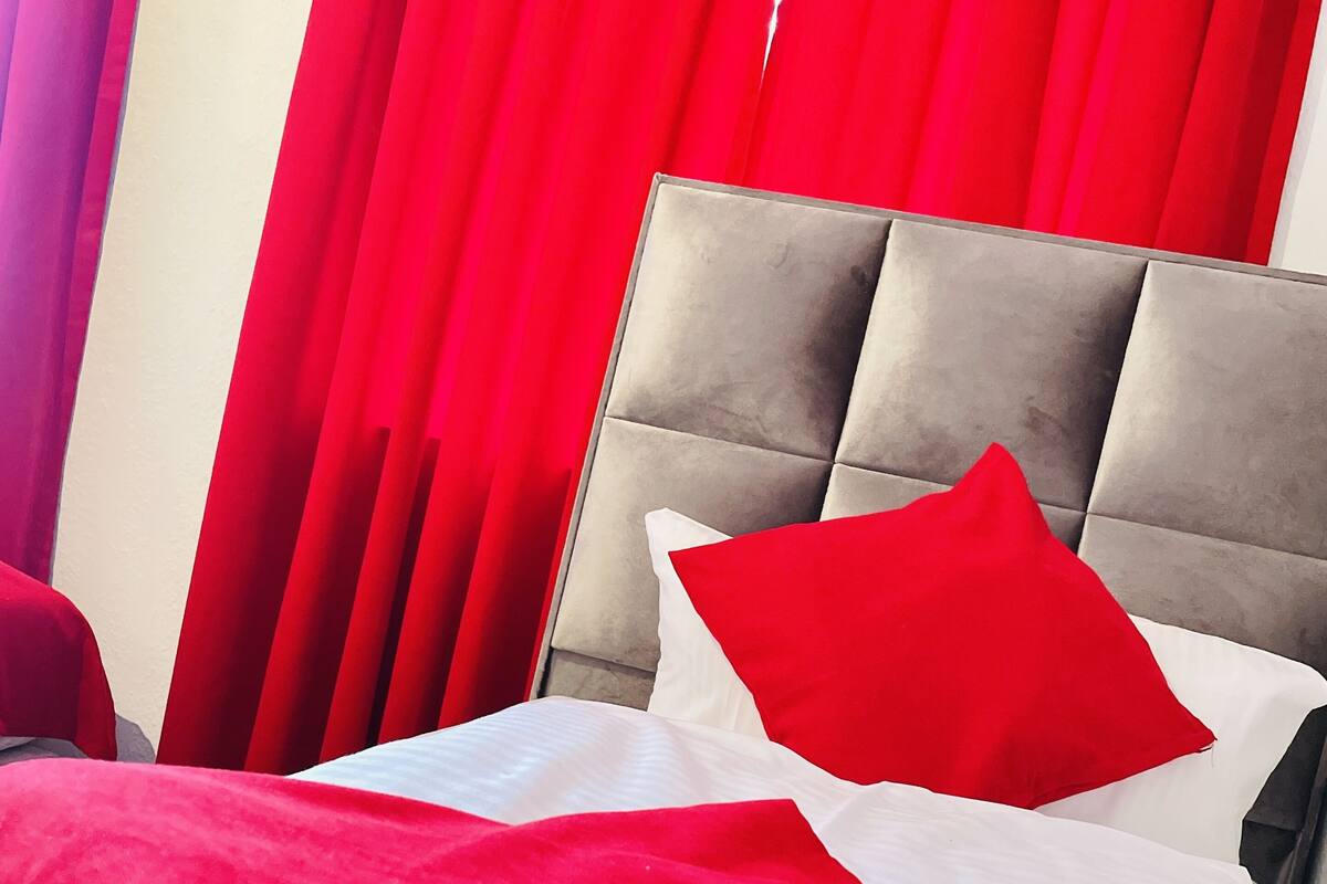 A bed with a red and white bedspread, a grey upholstered headboard, and red curtains in the background.